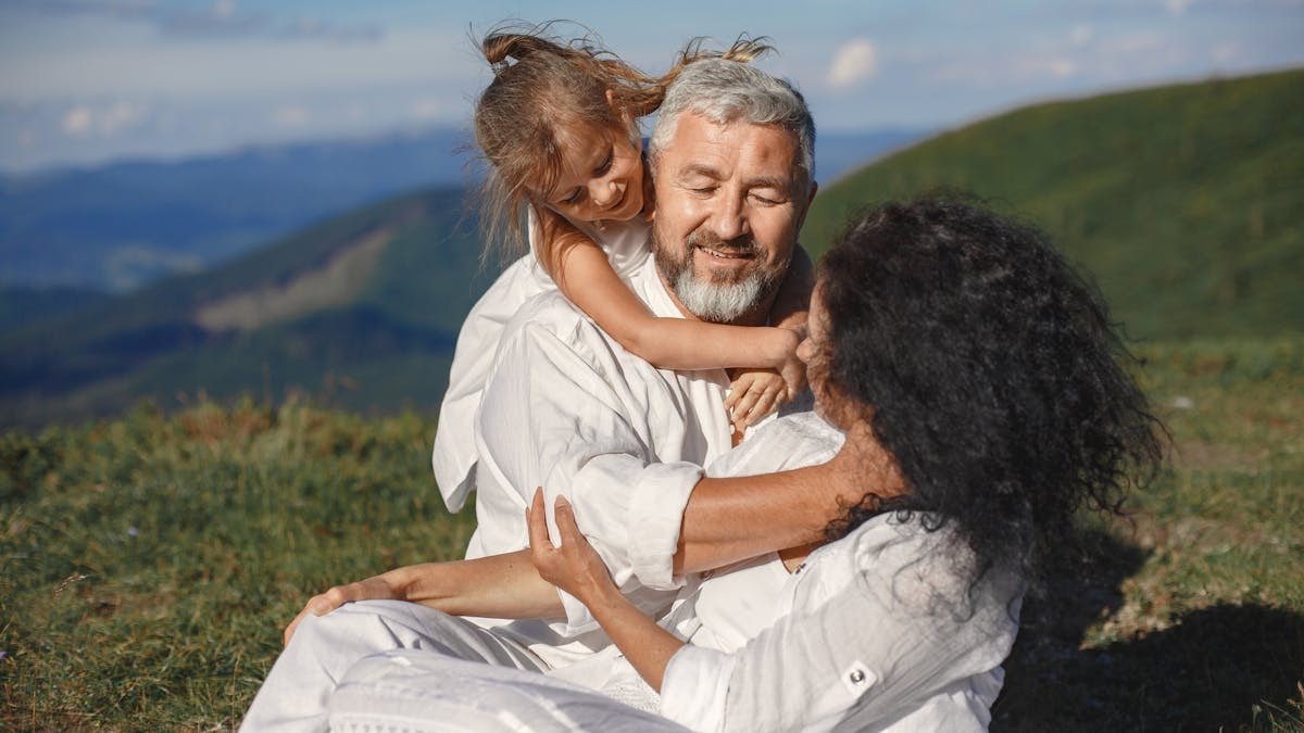 A joyful family moment outdoors, showing grandparents hugging their granddaughter under a sunny summer sky.
